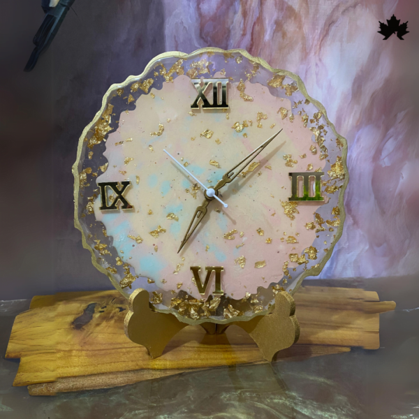 Alt text for the image: Blush Elegance Designer Table Clock by Fankaarlok, featuring a golden base supporting a round clock face with pastel pink and white hues, adorned with Roman numerals and gold flakes, positioned on a wood stand.