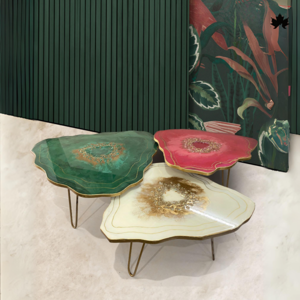 Three artistic resin bespoke agatet ables in varying shapes and vibrant colors of green, pink, and cream, each with unique golden accents and elegantly designed legs, presented against a marbled floor and an illustrated backdrop.